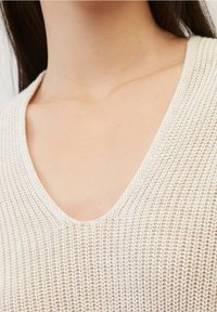 Jasno beżowy sweter z dzianiny z dekoltem w serek. Teksturalny wzór, luźny krój, prezentujący miękką, oddychającą tkaninę. Brak dodatkowych detali.
