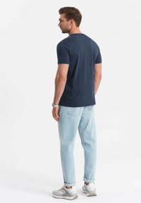 Námořnická modrá bavlněná tričko, krátký rukáv, kulatý výstřih. V kombinaci se světle modrými džínami a šedobílými teniskami. Pohled zezadu.