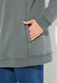 Felpa grigia con tasca laterale, polsini a costine e una texture morbida. Abbinata a jeans in denim blu.