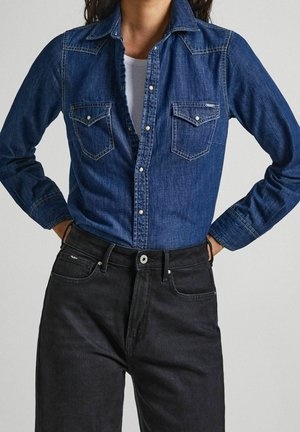 Person iført en blå denim skjorte med knapper over en hvid top, kombineret med højtaljede sorte jeans, hænderne hviler på hofterne.