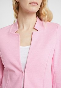 Žena s blond vlasy, která má na sobě zlaté náušnice ve tvaru kruhu, růžovou texturovanou bundu s otevřeným límcem a bílý top pod tím.