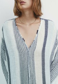 Blouse légère rayée blanc et marine, avec un décolleté en V et des manches amples aux bords effilochés. Le tissu texturé crée un contraste visuel.