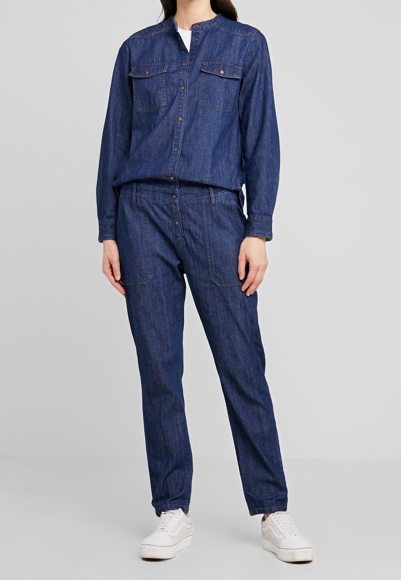 Femme portant une chemise bleue en denim à boutons avec poches poitrine et un pantalon en denim assorti avec poches plaquées, associée à des baskets blanches.