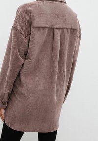 Chaqueta de camisa de pana marrón con un corte relajado, textura de canalé vertical, mangas largas y un yugo trasero para mayor detalle.