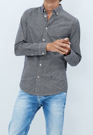 Homme portant une chemise sombre à motifs et manches longues boutonnée, ainsi qu'un jean bleu clair, les mains jointes à la taille devant un fond uni.