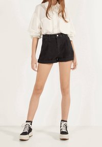 Camisa blanca de manga larga con mangas abullonadas y dos bolsillos en el pecho, combinada con pantalones cortos de mezclilla negros de cintura alta y zapatillas negras de caña alta.