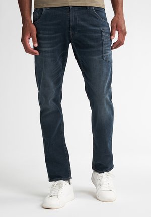 Man wearing dark blue straight-leg jeans with a side pocket and white lace-up sneakers on a white background.