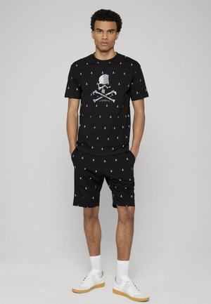 Mężczyzna stojący, ubrany w czarny t-shirt z wzorem czaszki i krótkie spodenki, z białymi trampkami i skarpetkami na jasnym tle.
