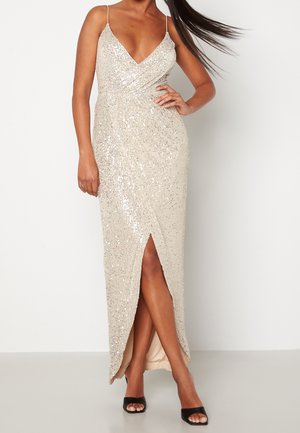 Kobieta ubrana w bezrękawnikową, srebrną suknię cekinową z dekoltem w kształcie litery V, z przednim rozcięciem i czarnymi sandałami z odkrytymi palcami, stojąca na jednolitym tle.