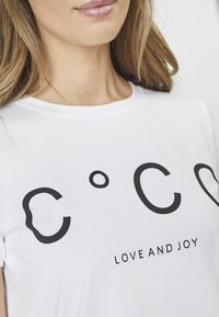 Camiseta de algodón blanca que presenta un texto negro en negrita: "C O C O" con un pequeño círculo y "AMOR Y ALEGRÍA" debajo, textura suave, escote redondo.