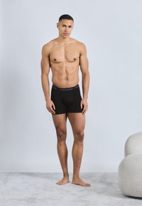 Μυώδης άνδρας που στέκεται ξυπόλητος σε χαλιμένο πάτωμα, φορώντας μαύρα εσώρουχα Tommy Hilfiger, μπροστά σε απλό λευκό φόντο.