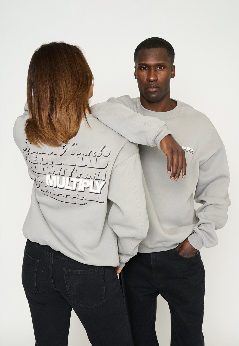 Grå sweatshirts med texturerade grafiska tryck på baksidan, visas på två modeller, tillsammans med svarta jeans. Rundad halsringning och lös passform.