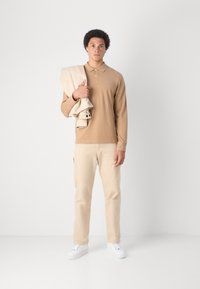 Polo de manga larga en tono beige, con cuello abotonado, combinado con pantalones beige claros y zapatillas blancas, sosteniendo una chaqueta de color crema.