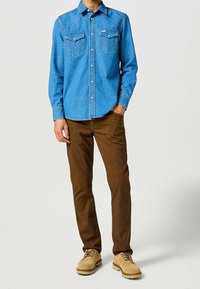 Camisa de mezclilla en azul claro con botones a presión, yugo puntiagudo y dos bolsillos en el pecho, combinada con pantalones marrones y zapatos beige.