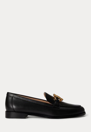 Mocassin en cuir noir avec un talon bas et un ornement en métal doré en forme de T-barre sur le dessus, présenté de profil sur un fond blanc.