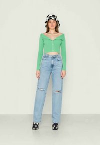 Top cropped verde com design off-the-shoulder, combinado com jeans rasgados de cintura alta em azul claro e sapatos pretos com padrão floral branco.
