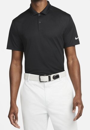 Mann trägt ein schwarzes kurzärmeliges Poloshirt mit weißem Nike-Logo auf dem Ärmel, weiße Hose und einen weißen gewebten Gürtel.