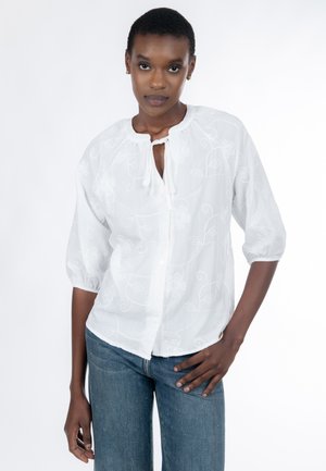 Hvid bluse med blomsterbroderi, rund halsudskæring med bindebånd, pufærmer og knapper foran. Sammen med blå jeans med pynt.