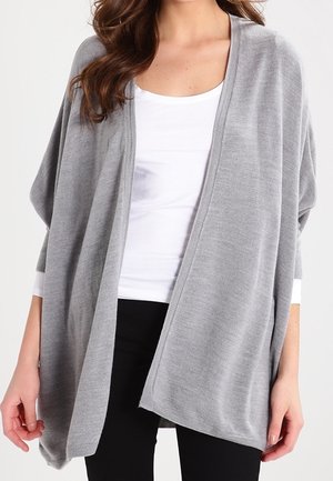 Femme portant un cardigan ample gris clair ouvert sur un haut blanc et un pantalon noir, avec des cheveux bruns ondulés visibles.