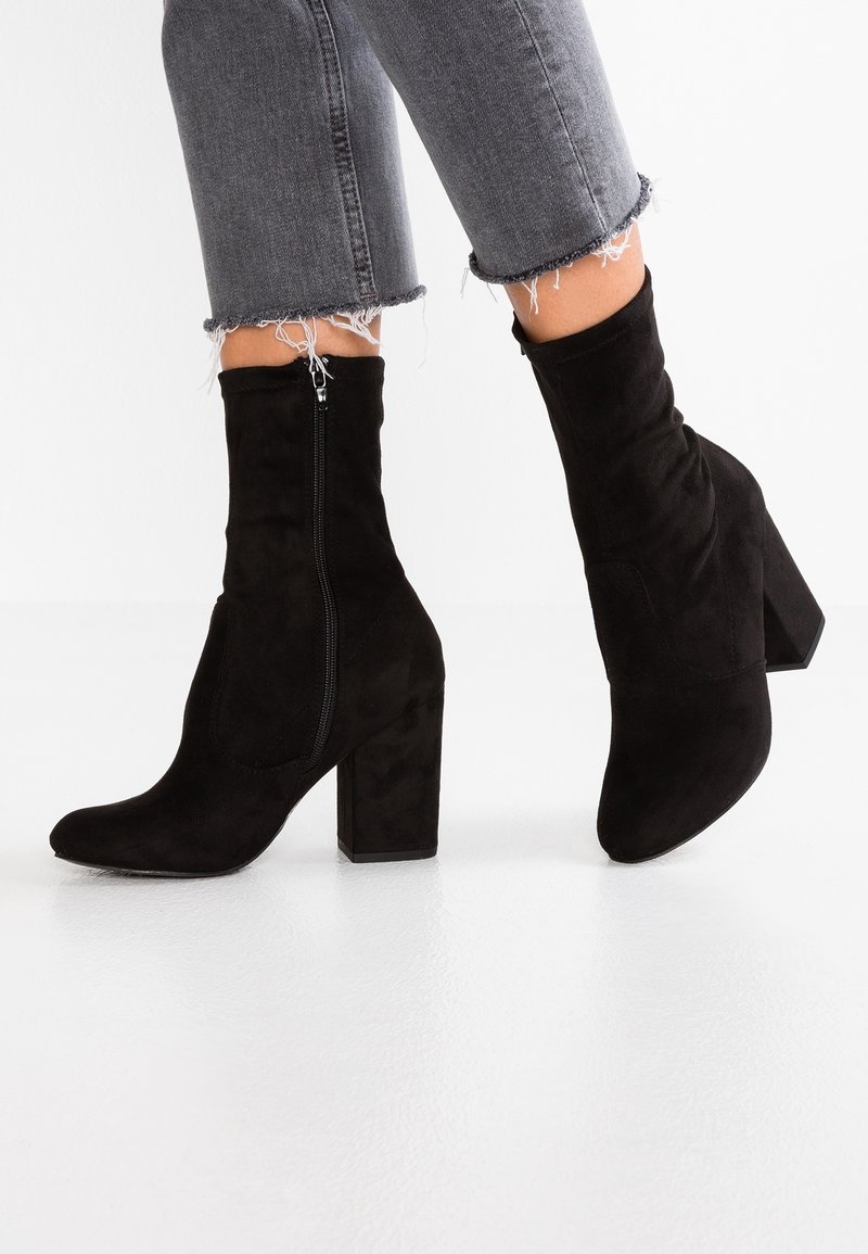 Personne portant des bottines en daim noir à talons épais et fermetures éclair latérales, associées à un jean gris à ourlets effilochés.