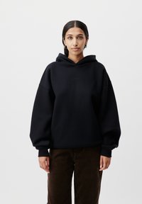 Felpa nera con cappuccio, dalla vestibilità oversize, con polsini a coste e logo ricamato sul petto. Indossata con pantaloni in velluto a coste marroni.