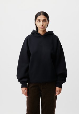 Sweatshirt à capuche noir avec coupe oversize, poignets côtelés et logo brodé sur la poitrine. Porté avec un pantalon en velours côtelé marron.