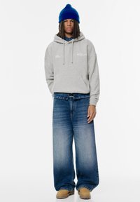 Grå huvtröja med framlapp, blå mössa, vida blå jeans och bruna stövlar. Enkelt mönster med synlig varumärkestext på huvtröjan.