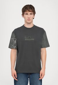Tamsiai pilkas medvilnės marškinėliai su puoštomis trumpomis rankovėmis iš tekstūrinio audinio. Centre esantis logotipas neryškiai žalia spalva: „EA7 Emporio Armani“. Laisvas siluetas.