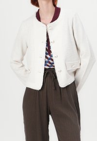 Biała teksturowana kurtka z okrągłym dekoltem i postrzępionymi krawędziami, zapięta jasnymi guzikami, noszona na wzorzystą koszulę i ciemne spodnie.