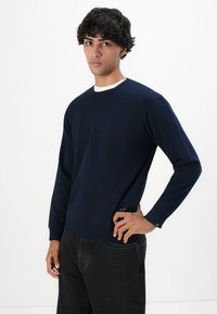 Joven con cabello rizado y oscuro que lleva un sudadera azul marino sobre una camisa blanca y jeans negros, con la mano descansando en la cadera.