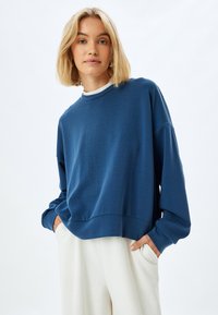 Felpa di cotone blu con maniche lunghe e scollo rotondo. Design cropped e texture liscia, abbinata a pantaloni a gamba larga beige chiaro.