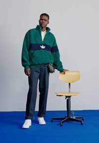 Pull en laine verte duveteux avec une rayure marine, logo blanc ; pantalon sombre, baskets blanches. Chaise en bois à côté du modèle, sur un fond bleu.