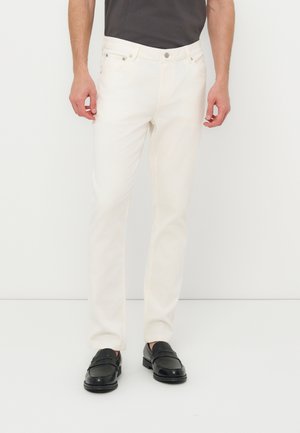 Άνδρας που φορά στενά άσπρα τζιν και μαύρα δερμάτινα μοκασίνια, όρθιος μπροστά σε λευκό φόντο.