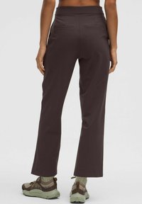 Sötétbarna, rugalmas nadrág magas derékkal és enyhén bővülő szárakkal a bokánál. Hátul zsebekkel rendelkezik és világos sportcipőkkel van párosítva.