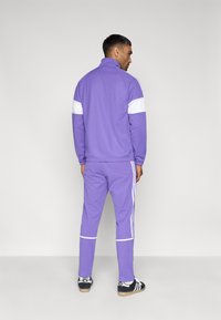 Μωβ φόρμα με λευκές λεπτομέρειες, που περιλαμβάνει μπουφάν με φερμουάρ στο γιακά και ταιριαστό παντελόνι με ρίγες στα πλάγια. Το ύφασμα φαίνεται απαλό και λείο.