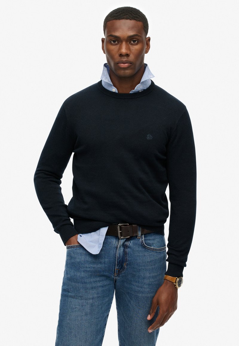 Marineblauer Strickpullover mit Rundhalsausschnitt, über einem hellblauen Hemd mit Kragen getragen. Dazu dunkelblaue Jeans und ein brauer Ledergürtel.