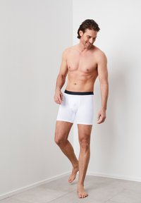 Weiße Boxershorts mit einem dunklen Bund, hergestellt aus einem glatten, dehnbaren Stoff, mit einem enganliegenden Schnitt und einer Länge bis zur Mitte des Oberschenkels.