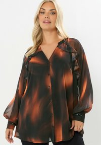 Blusa semitransparente negra y naranja con cuello con volantes, mangas largas y cierre de botón. Detalles finales con puños con tachuelas.