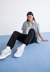 Lys grå kortærmet skjorte over en hvid t-shirt, parret med sorte sweatpants og hvide sneakers med grå accenter. Sidder på en blå overflade.
