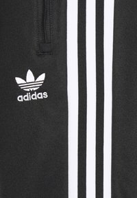 Černé sportovní kalhoty s třemi bílými vertikálními pruhy. Obsahují vyšitý bílý logo Adidas. Materiál vypadá texturovaně a odolně.