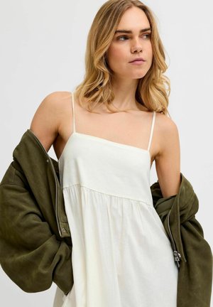 Szőke nő fehér ujjatlan ruhában túlméretes olívazöld dzsekit visel lehúzva a válláról, jobbra néz egy egyszínű háttér előtt.