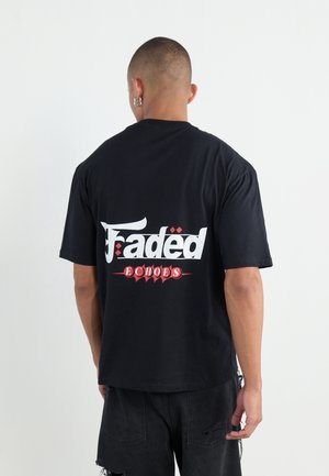 Homme aux cheveux courts et boucles d’oreilles en anneau porte un T-shirt noir avec le texte « Faded Echoes » en blanc et rouge dans le dos, assorti à un short noir usé.