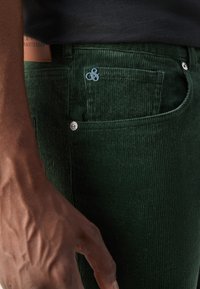 Ådrad hand vilande nära fickan på mörkgröna corduroybyxor med metalknappdetaljer och en liten silverlogotypnål.