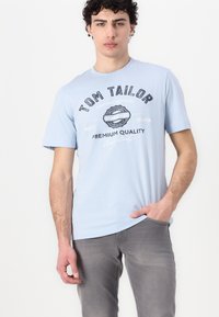 Tricou din bumbac de culoare albastru deschis, cu un print grafic ce include textul "TOM TAILOR" și un design de insignă circulară. Mâneci scurte și guler de tip crew neck.