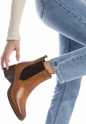 Botas de cuero marrón hasta el tobillo con paneles laterales elásticos acanalados, punta redondeada y un tacón bajo y apilado, combinadas con jeans de color azul claro.