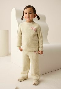 Cream-colored knit sweater and pants set with pink "NYC" lettering, featuring ribbed cuffs and a thick, soft texture. Complemented by light sneakers.