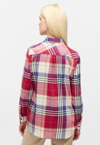 Donna con lunghi capelli biondi che indossa una camicia a quadri larga rossa, blu e beige, vista di spalle su uno sfondo chiaro e semplice.