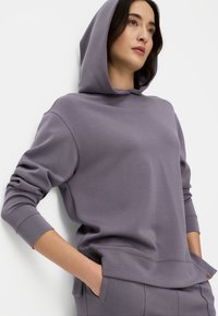 Paarse hoodie met een losse pasvorm, lange mouwen en een voorzak. Gladde textuur met subtiele stikdetails.