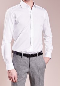 Camisa blanca de manga larga con cuello clásico, botones al frente y tejido liso, combinada con pantalones grises texturizados y cinturón negro.