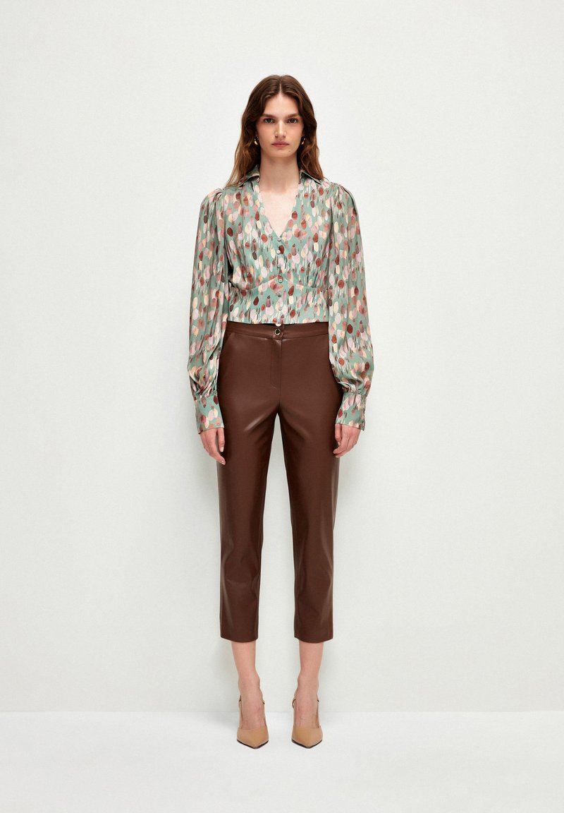 Blusa a maniche lunghe, stampata in verde con accenti rosa e beige, abbinata a pantaloni in ecopelle color marrone, tagliati e a tacco nudo.
